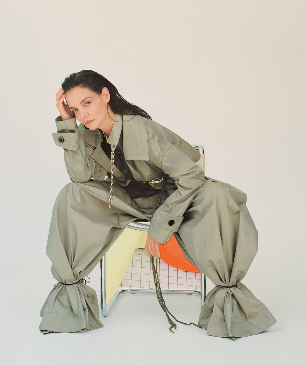 Woman in an olive green utility jumpsuit with cinched ankles, seated on a modern metal frame chair against a plain white background