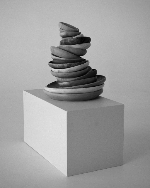 Monochrome studio shot: stacked wooden bowls create a sculptural form on a white cube against a neutral background, artful display.