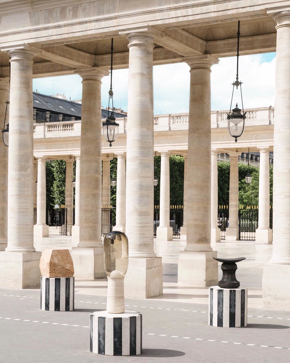 Outdoor art installation with sculptures on striped pedestals in an arcade with columns and hanging lanterns, set on a paved surface.