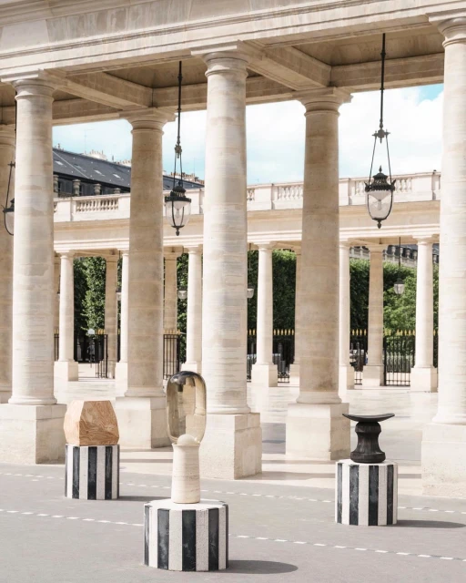 Outdoor art installation with sculptures on striped pedestals in an arcade with columns and hanging lanterns, set on a paved surface.