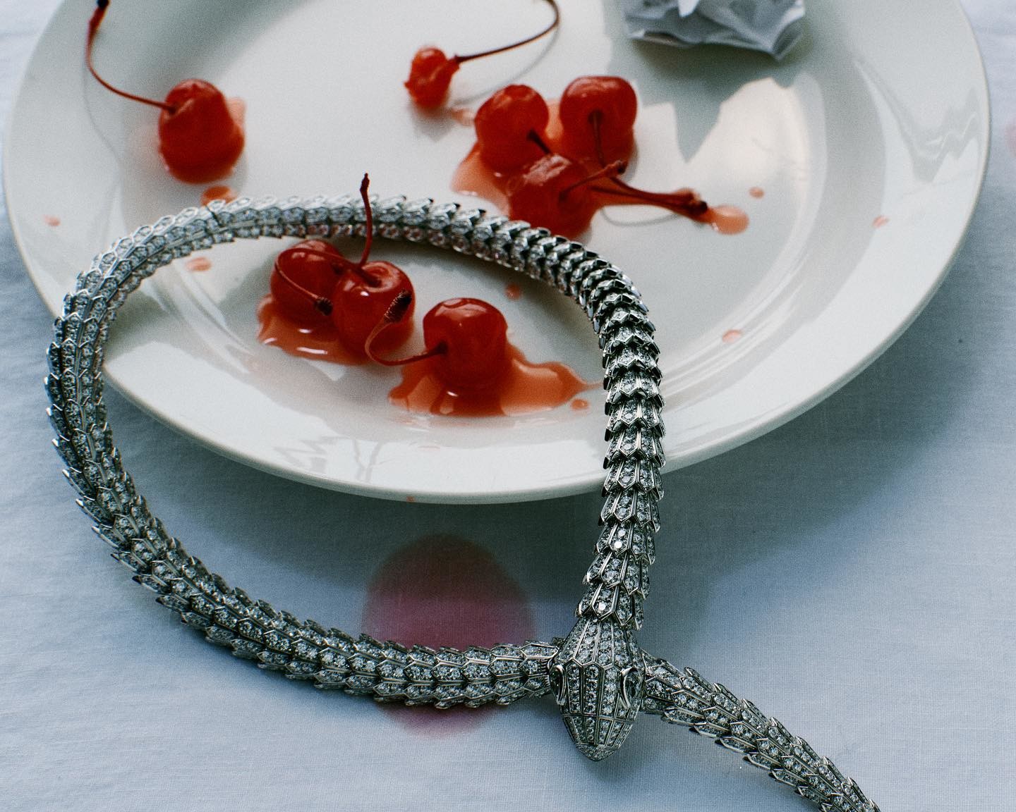 Elegant diamond snake necklace encircles a plate of red cherries with stems, accompanied by a crumpled white paper ball.