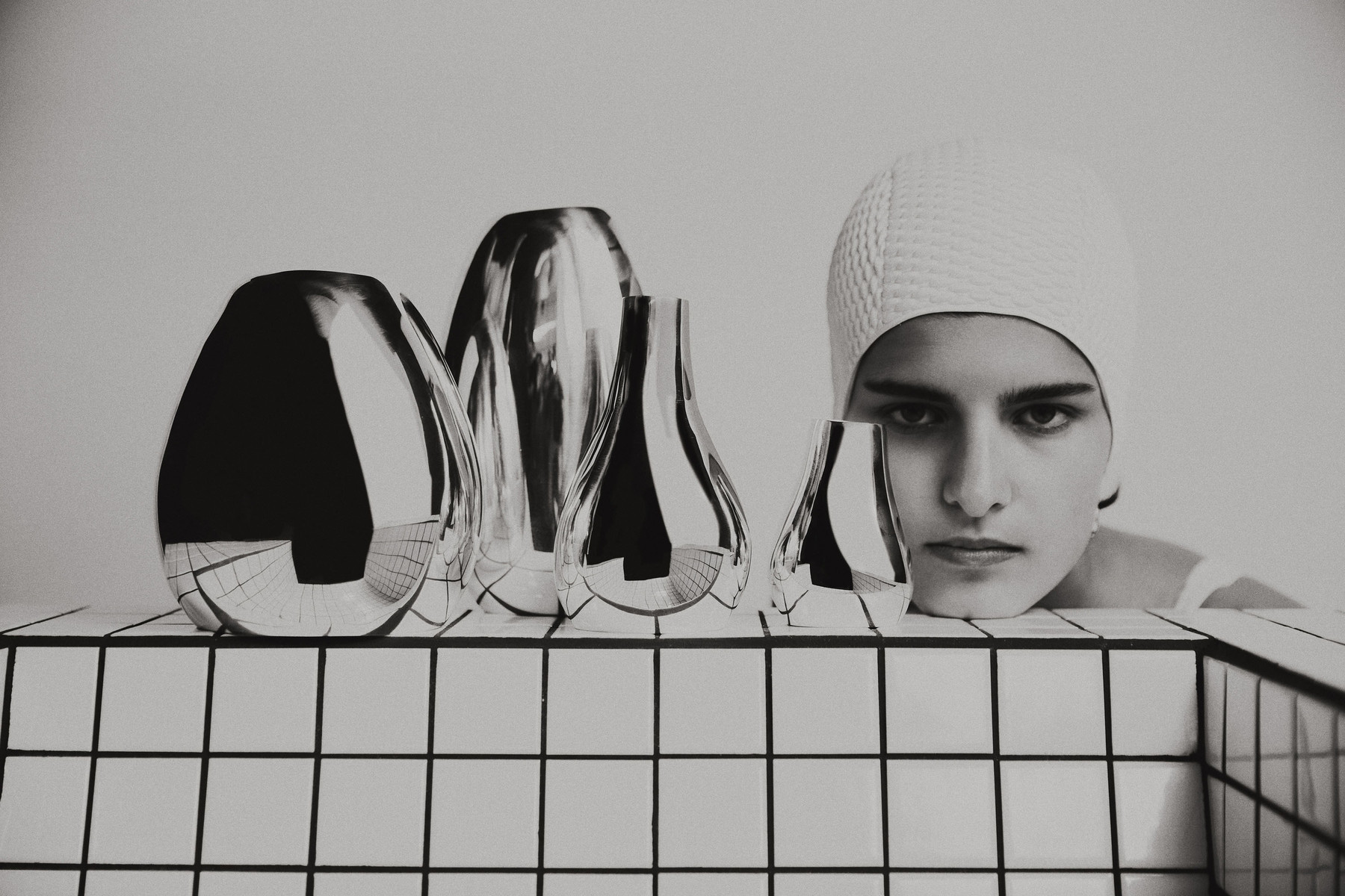 Monochrome studio shot features a woman in a swim cap posing next to shiny, sculptural vases against a white tiled backdrop.