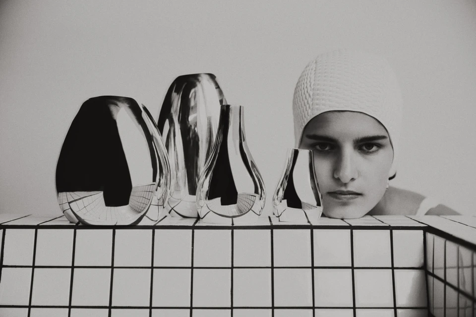 Monochrome studio shot features a woman in a swim cap posing next to shiny, sculptural vases against a white tiled backdrop.