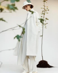 Model in white textured coat, bucket hat, and trousers standing on a white background with a small potted tree in the foreground.