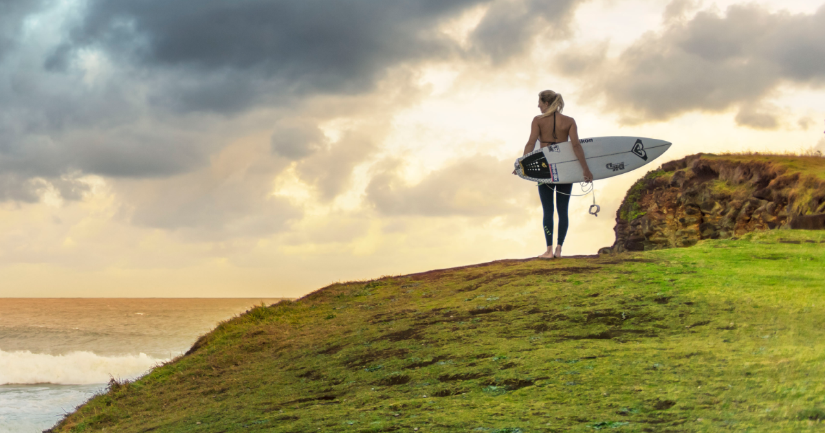 A female surfer stands on a grassy cliff overlooking the ocean with her surfboard at sunset under a cloudy sky.