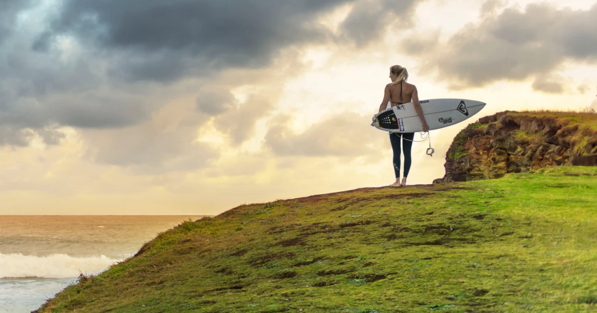 A female surfer stands on a grassy cliff overlooking the ocean with her surfboard at sunset under a cloudy sky.