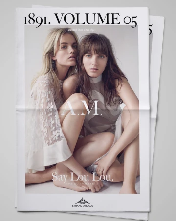 Two women pose on a newspaper advertisement for Say Lou Lou. They wear sheer blouses and pose indoors with neutral lighting.