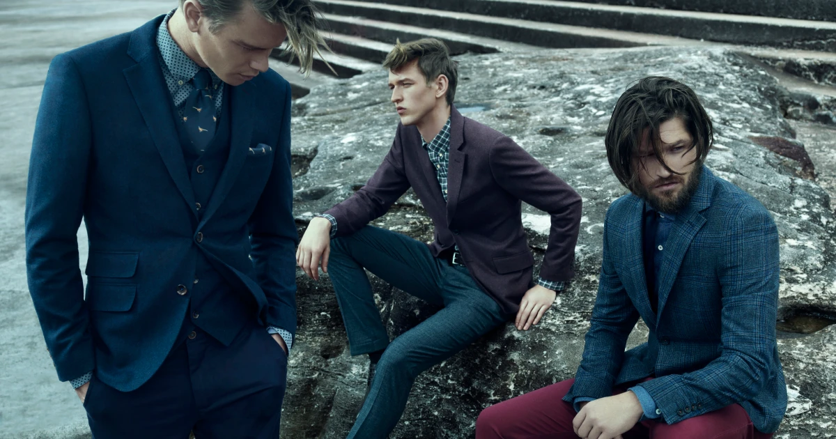Three male fashion models in suits pose outdoors against a textured stone wall wearing suits in blue and burgundy hues.