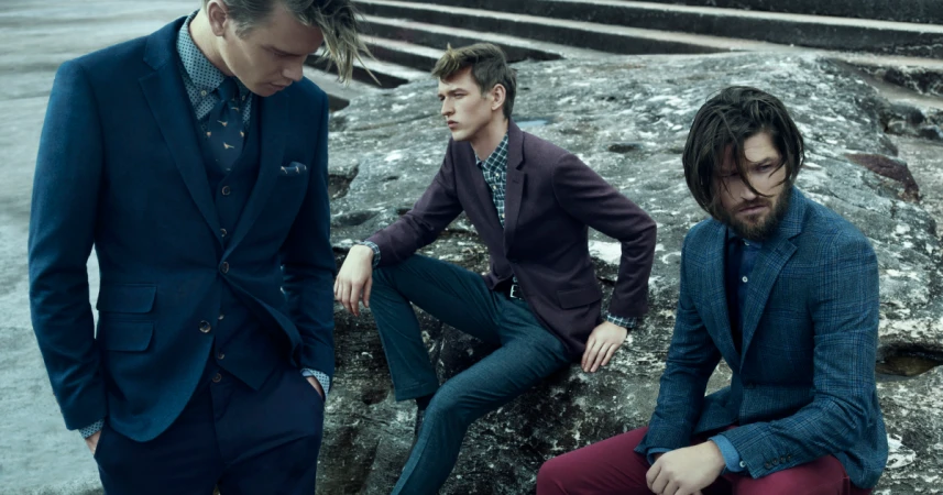 Three male fashion models in suits pose outdoors against a textured stone wall wearing suits in blue and burgundy hues.