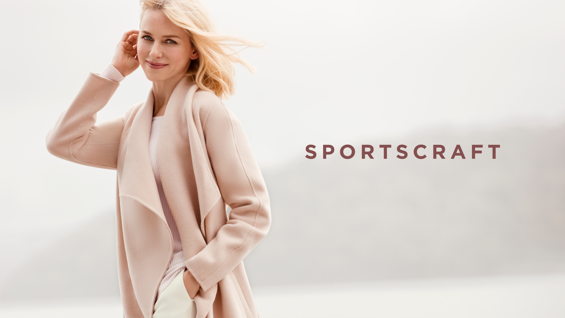 Blonde woman in pink coat and white pants posing outdoors for Sportscraft fashion brand advertisement with soft lighting and foggy background.