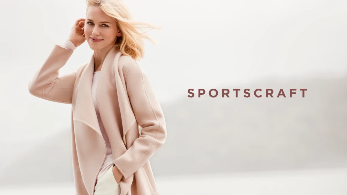 Blonde woman in pink coat and white pants posing outdoors for Sportscraft fashion brand advertisement with soft lighting and foggy background.
