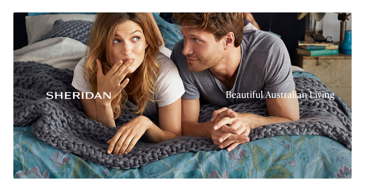 Attractive couple lounges in bed on a floral-patterned duvet beneath a grey knitted blanket, with natural light and a cozy ambiance.