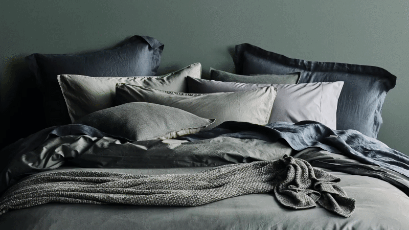 A neatly made bed with an assortment of soft pillows and a folded knit blanket, set against a muted green wall in a peaceful setting.