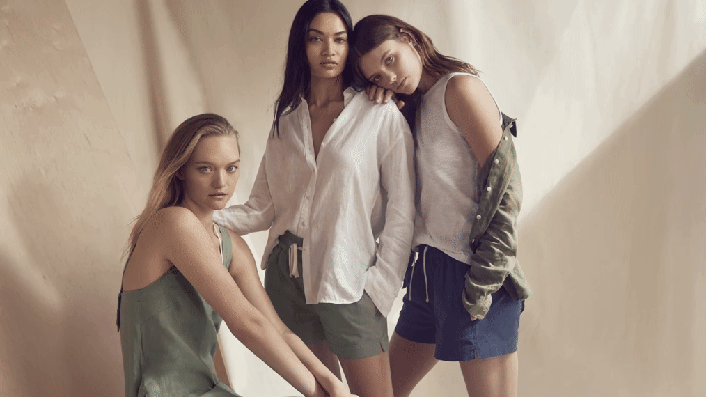 Three female fashion models pose indoors wearing casual shirts and shorts in a studio with soft lighting and a neutral background.