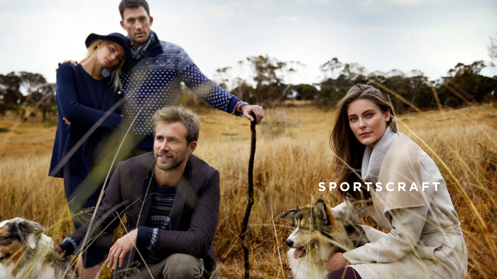 Outdoor fashion shoot with four models, two dogs, and tall grass in a field, showcasing autumn clothing and style.