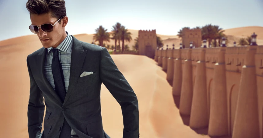 Man wearing a dark suit, plaid shirt, and aviator sunglasses outdoors against a desert fortress backdrop.