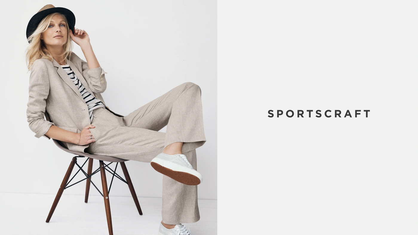 Blonde woman wearing beige linen suit jacket and trousers seated on a mid-century modern chair against a white background