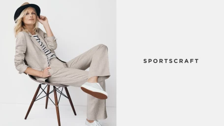 Blonde woman wearing beige linen suit jacket and trousers seated on a mid-century modern chair against a white background