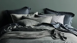 A neatly made bed with layered dark grey and neutral linen pillows and blankets against an olive green wall.