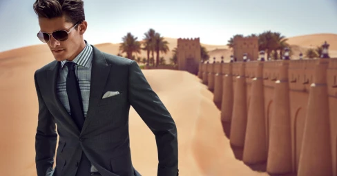 Man wearing a dark suit, plaid shirt, and aviator sunglasses outdoors against a desert fortress backdrop.