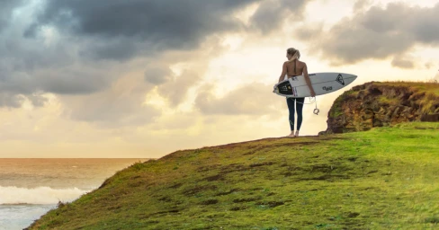 A blonde woman holding a surfboard stands on a grassy clifftop overlooking the ocean under a dramatic cloudy sky.