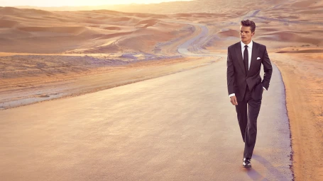 Man in dark suit and tie walking along an asphalt road winding through a vast sandy desert landscape.
