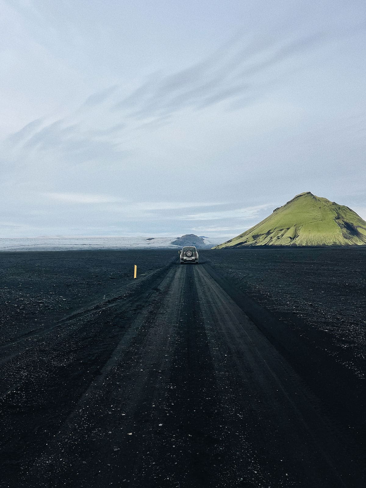 Vehicle travels down a gravel road across a black sand plain in Iceland, with mountains and cloudy sky visible in the distance.