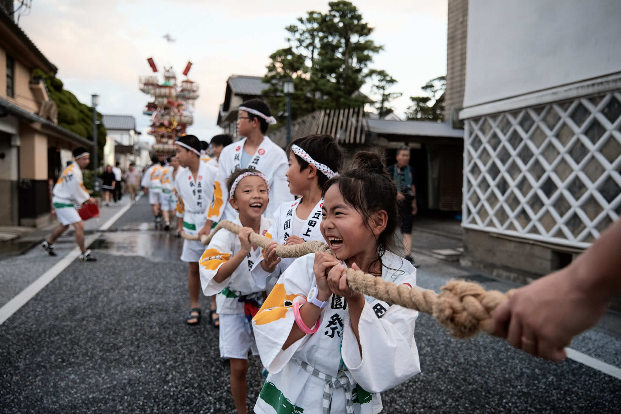 Children in traditional Japanese clothing pulling a rope down a street, participating in a festival with a decorative tower.