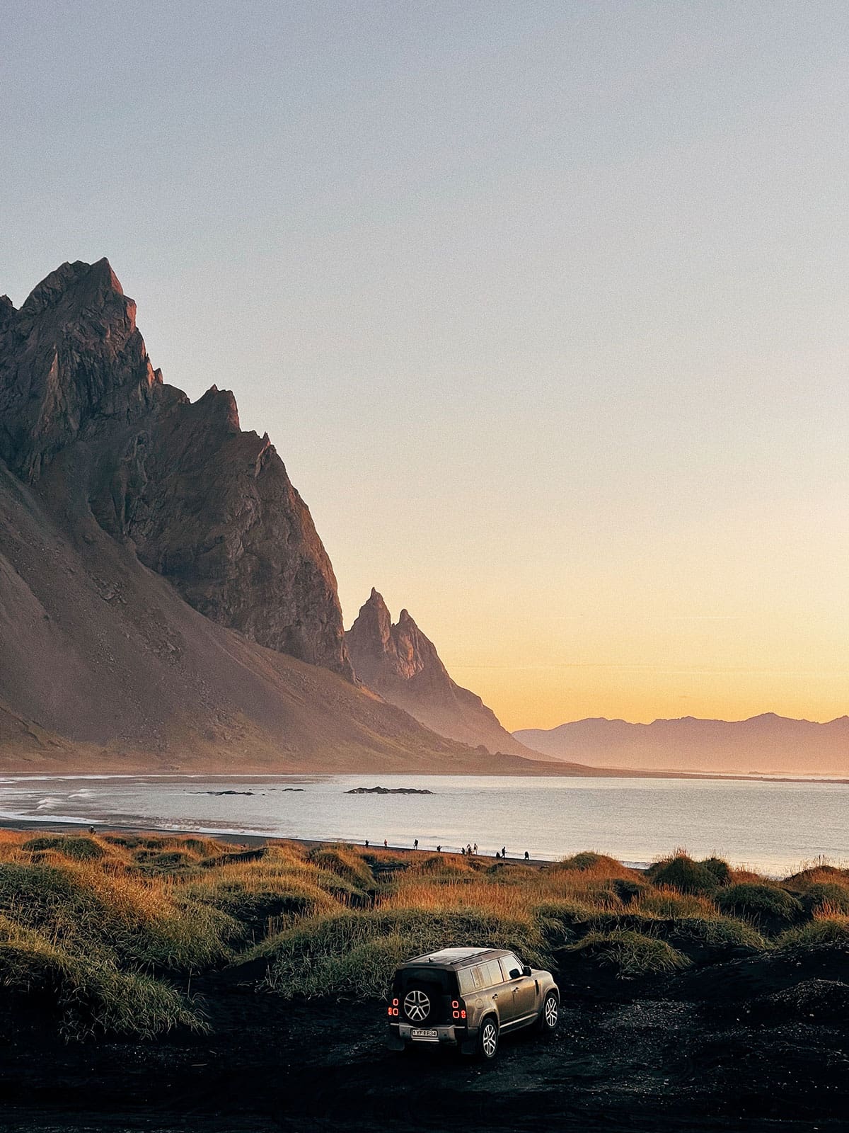 A scenic view of a silver SUV on a black sand road, with a mountain range and the ocean in the background during golden hour.