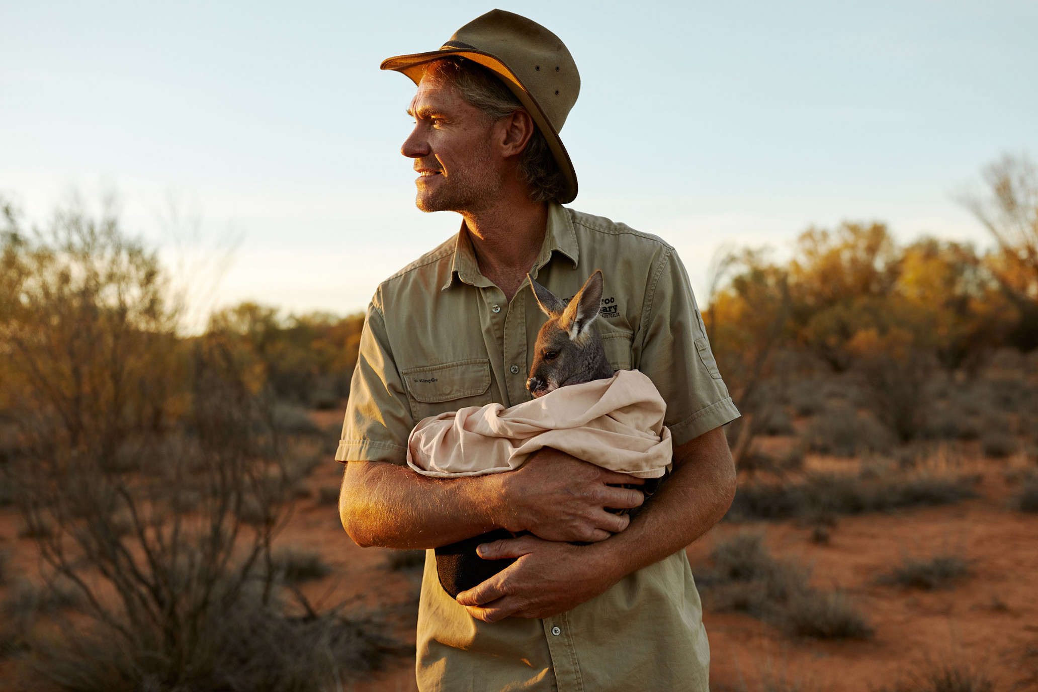 Man in khaki shirt and hat holds a baby kangaroo wrapped in a blanket, standing in a desert landscape at sunset.