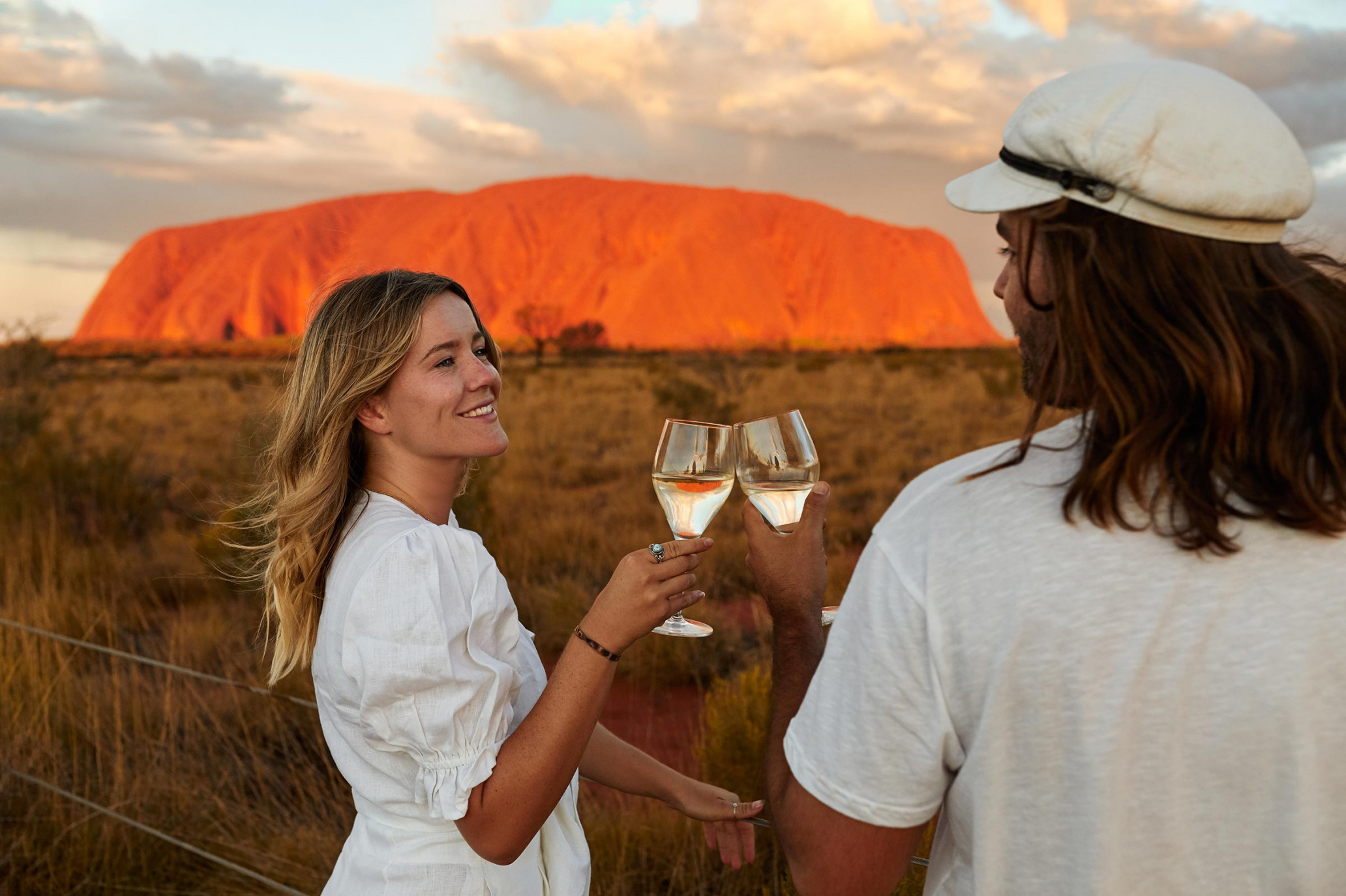 Couple toasting wine glasses in front of Uluru at sunset. The sky glows with warm colors as they celebrate in the Australian outback.