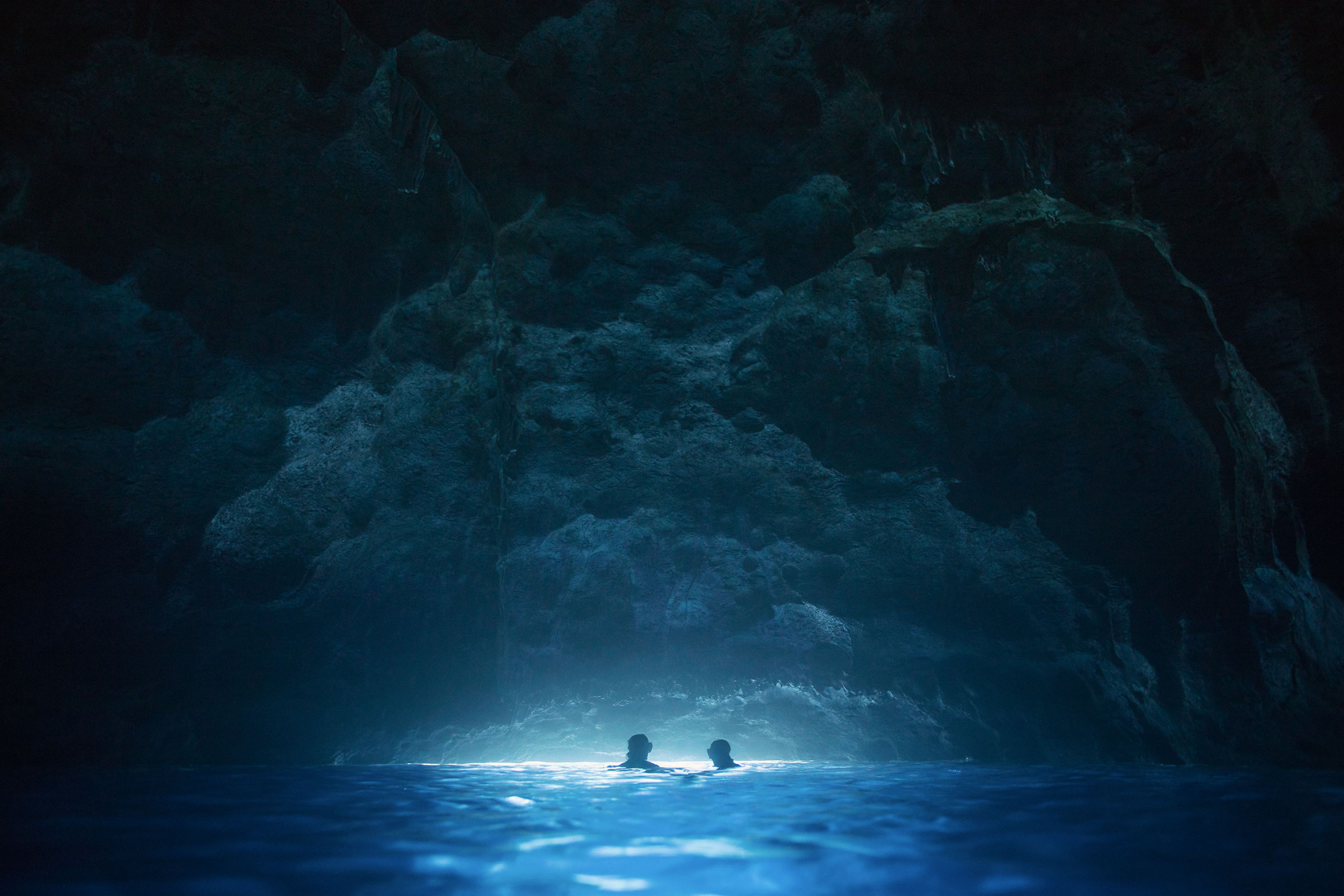 Two people swim in a dark cave with illuminated turquoise water. The rock formations provide an enclosed, cavernous setting.