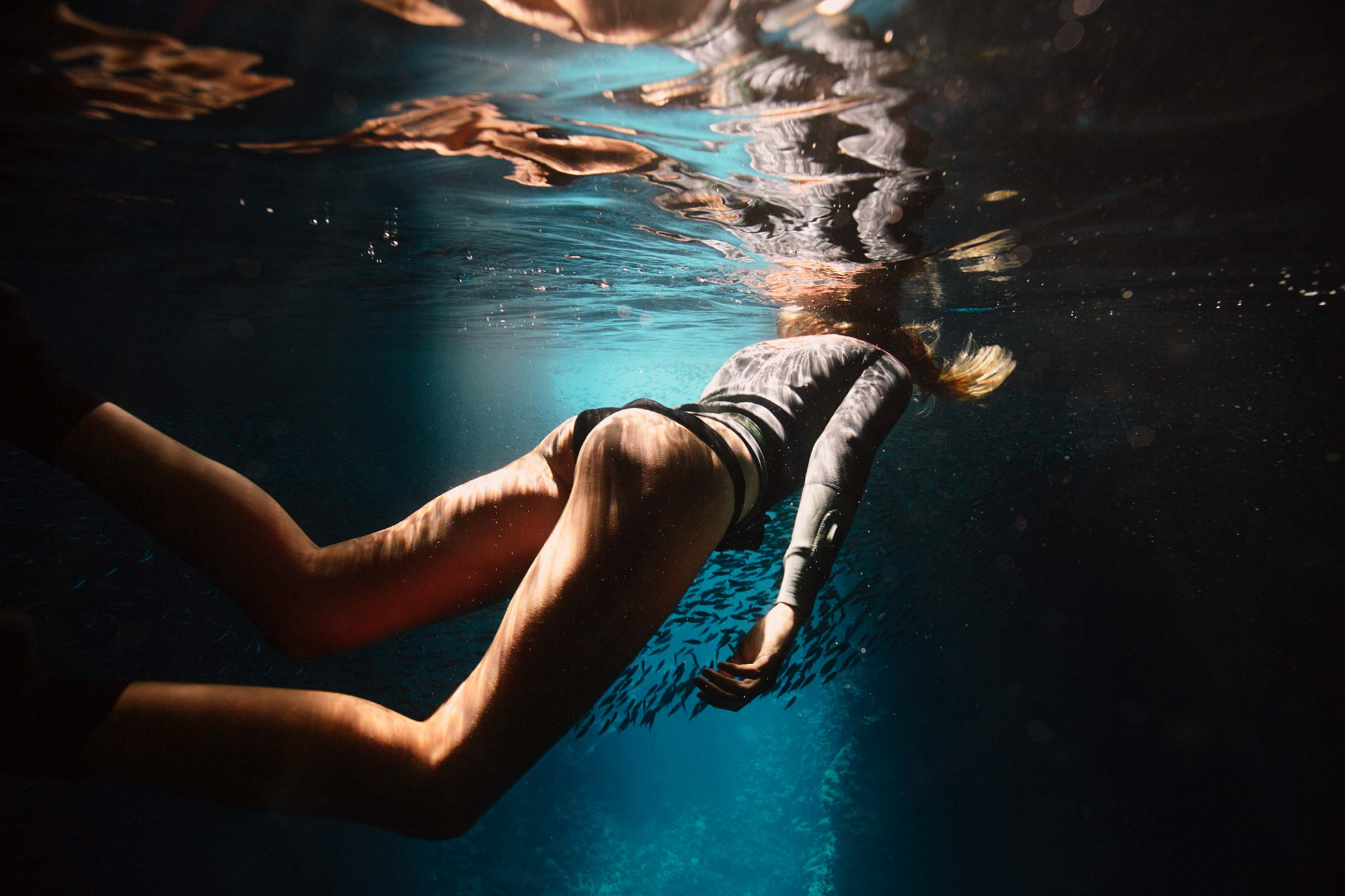 Underwater shot of a blonde woman in a wetsuit swimming, with a school of fish in the background, capturing an athletic moment.