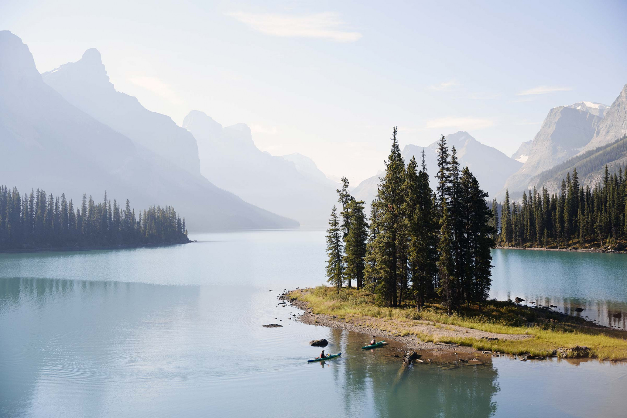 Scenic view of a lake with islands and kayakers, surrounded by forested mountains under a bright sky, capturing the serenity of nature.