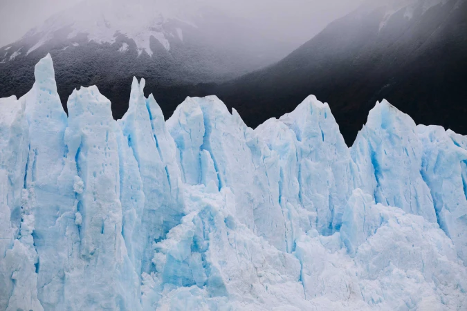 Jagged bright blue glacial ice wall against dark snow-dusted mountains under an overcast sky.