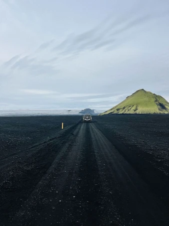 Car driving away on a dark volcanic black sand road beneath an overcast sky with a prominent green mountain in the distance.