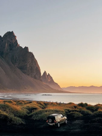An off-road vehicle parked on dark terrain before a tranquil coastal body of water backed by jagged mountains at sunset.