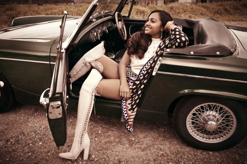 A smiling woman in a vintage convertible car wearing high fashion boots and a polka-dot top, with a gravel road and grassy field.