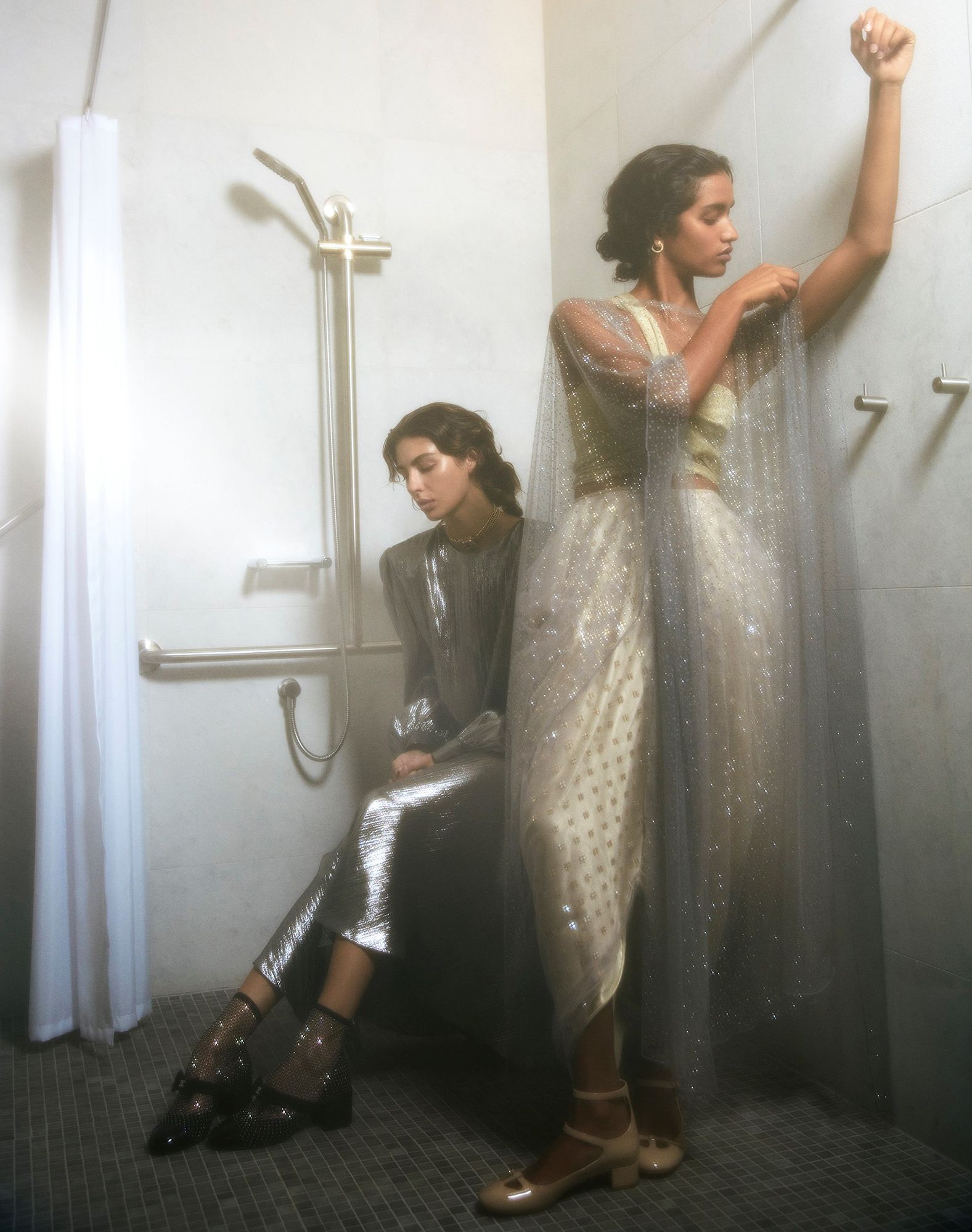 Two women in sparkling gowns pose in a tiled room. One sits, the other leans against the wall with a shower fixture behind her.