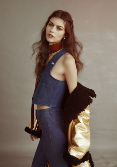 Woman wearing denim vest and pants with a gold metallic jacket draped over her shoulder against a neutral grey backdrop.