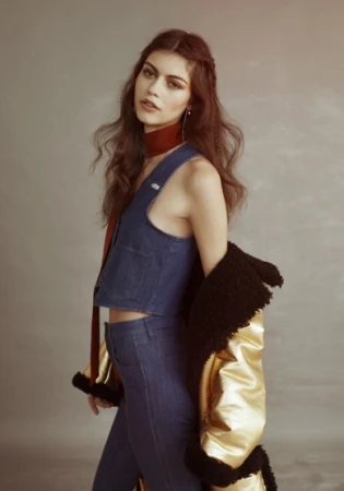 Woman wearing denim vest and pants with a gold metallic jacket draped over her shoulder against a neutral grey backdrop.