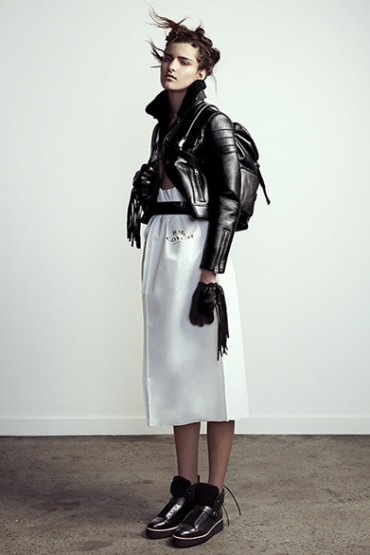 Full-length portrait of a female fashion model wearing a black leather jacket and backpack, a white skirt, and black boots in studio.