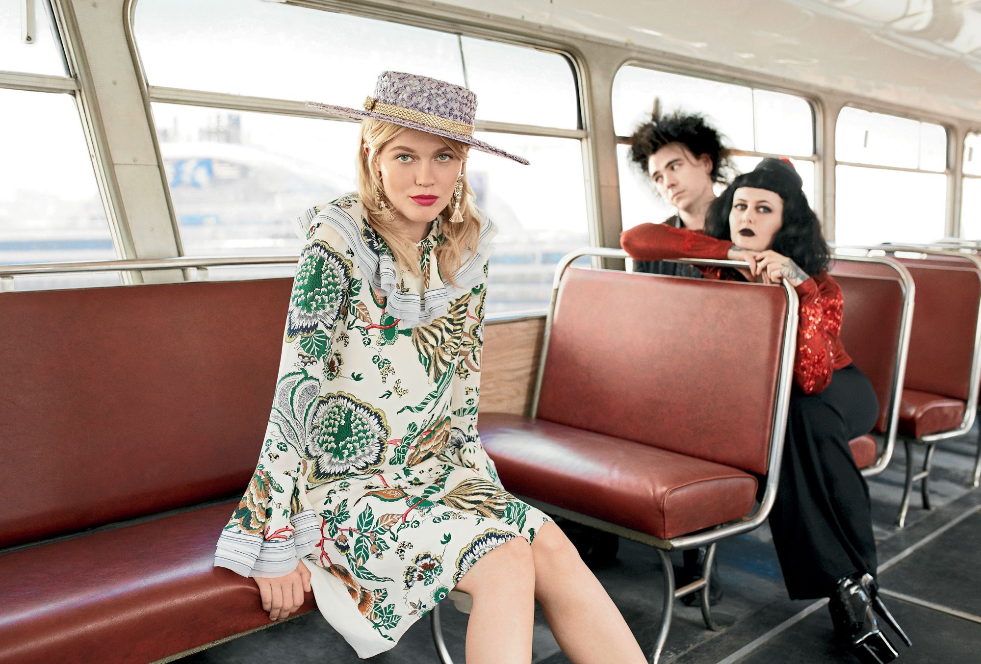Fashion shot of a woman in a patterned dress and hat sitting on a vintage bus with two people visible in the background.