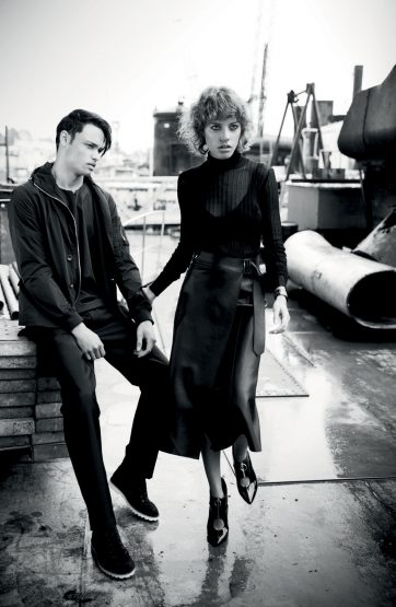 Black-and-white fashion shot of an adult male sitting and an adult female standing in an industrial outdoor setting, wearing dark clothing.