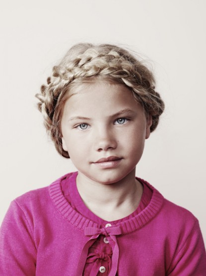 Close-up of a blonde girl with a braided crown hairstyle. She is wearing a pink sweater, and the background is a plain light color.