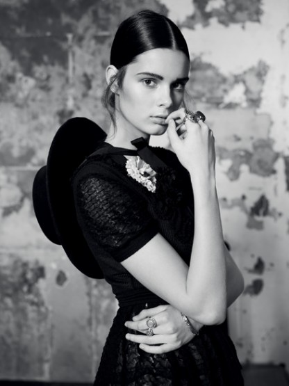 Portrait of a stylish woman in black dress and hat with vintage jewelry, posing against a textured wall in a black-and-white editorial.