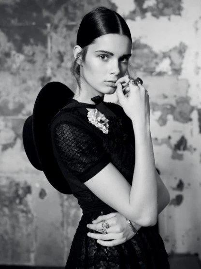 Portrait of a stylish woman in black dress and hat with vintage jewelry, posing against a textured wall in a black-and-white editorial.