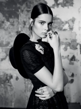 Portrait of a stylish woman in black dress and hat with vintage jewelry, posing against a textured wall in a black-and-white editorial.