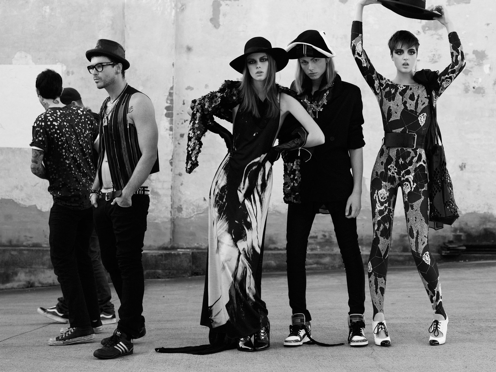 Black-and-white fashion editorial shot of four people in hats and stylish outfits, standing against a neutral wall in an urban setting.