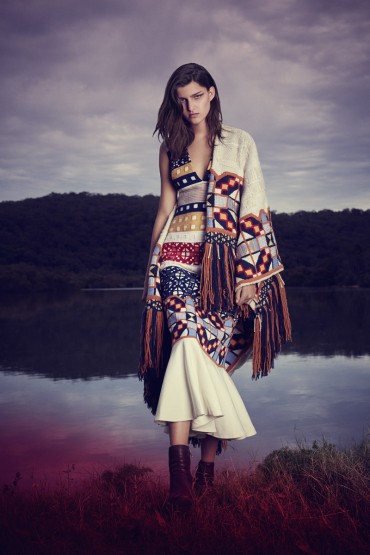 A female model stands in a patterned dress and fringed poncho in an outdoor setting, with a lake and hill visible in the background.